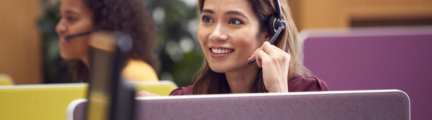 Mature Businesswoman Wearing Phone Headset Talking To Caller In Busy Customer Services Centre